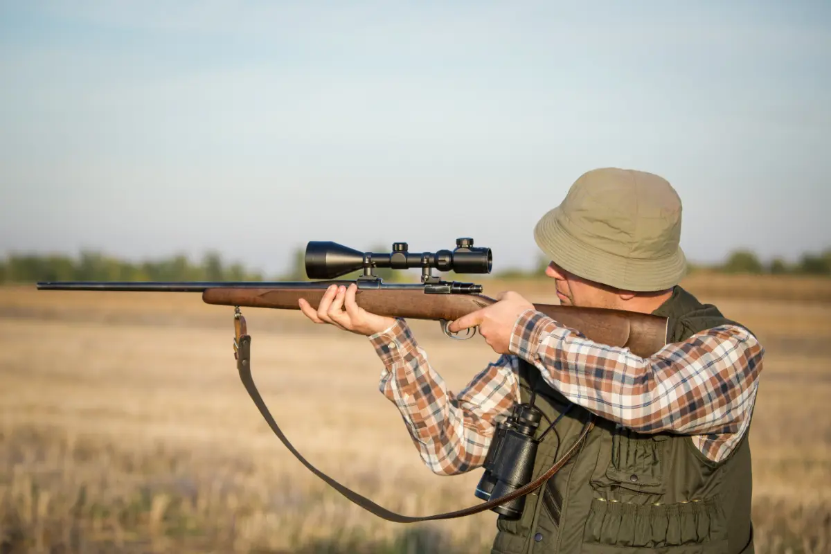 Ein Jäger zielt mit einem Gewehr, das mit einem Zielfernrohr ausgestattet ist, auf ein Ziel in einem offenen Feld, während er einen olivgrünen Hut und eine Weste trägt.